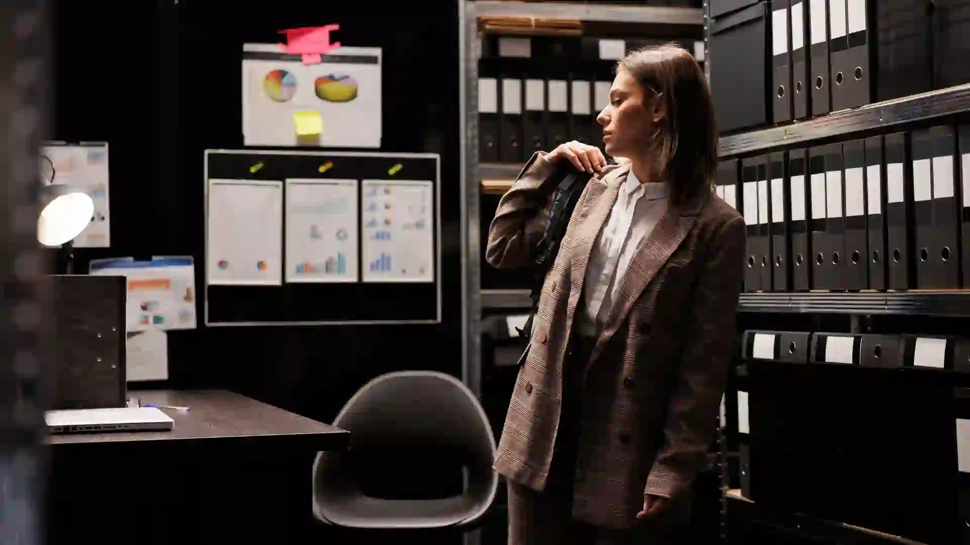 Corrections department staff member standing in an evidence archive room, surrounded by shelves of case files and binders, reviewing investigative charts and reports on the wall.
