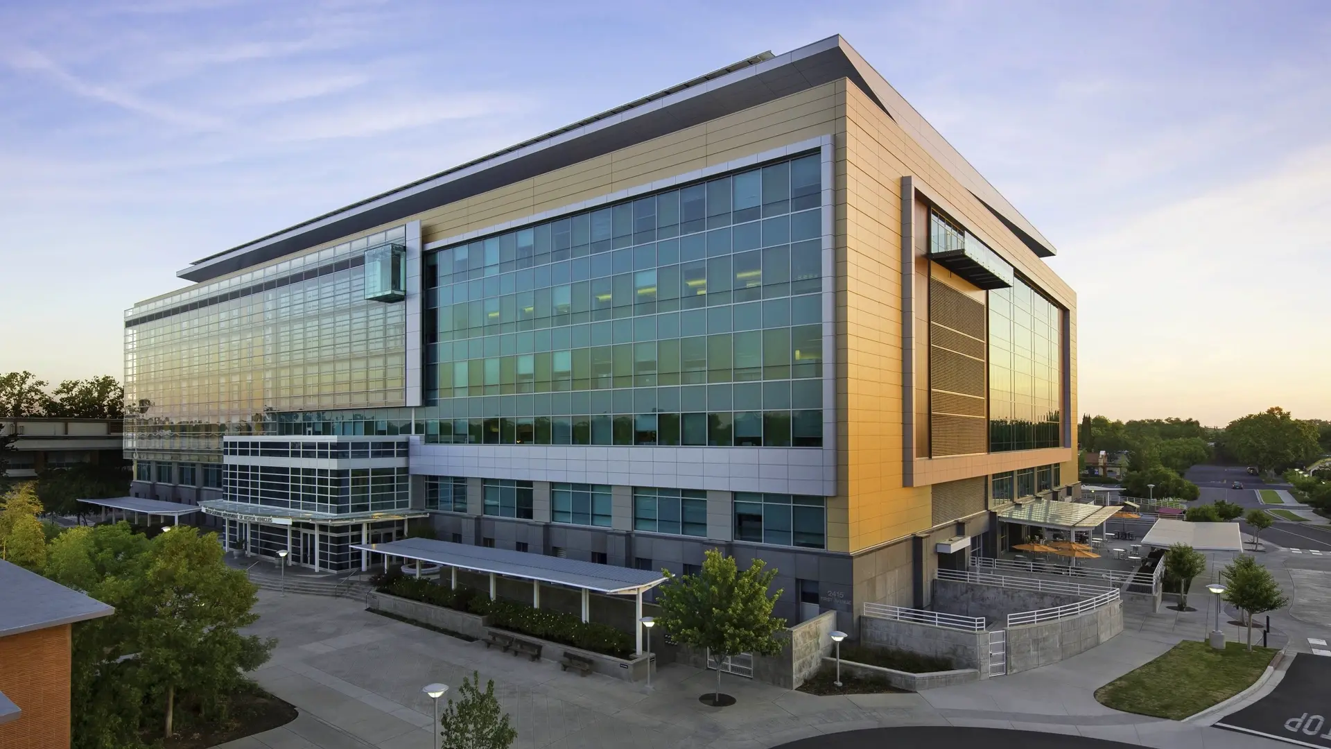 Exterior view of the California DMV headquarters, a modern building with large glass windows reflecting the sunset, surrounded by trees and open pathways.