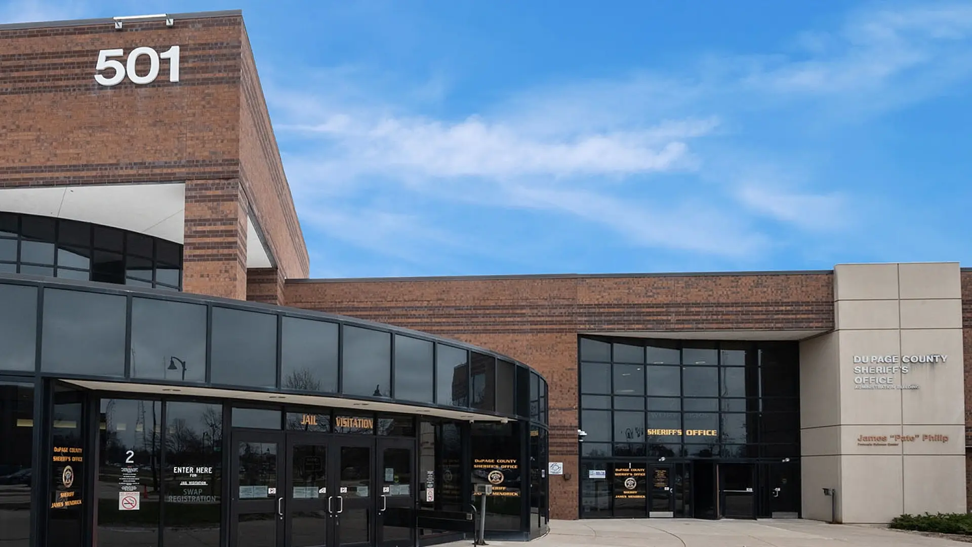 Exterior view of DuPage County Sheriff's Office building in Illinois, showcasing its modern architectural design with large glass panels and brickwork under a clear blue sky