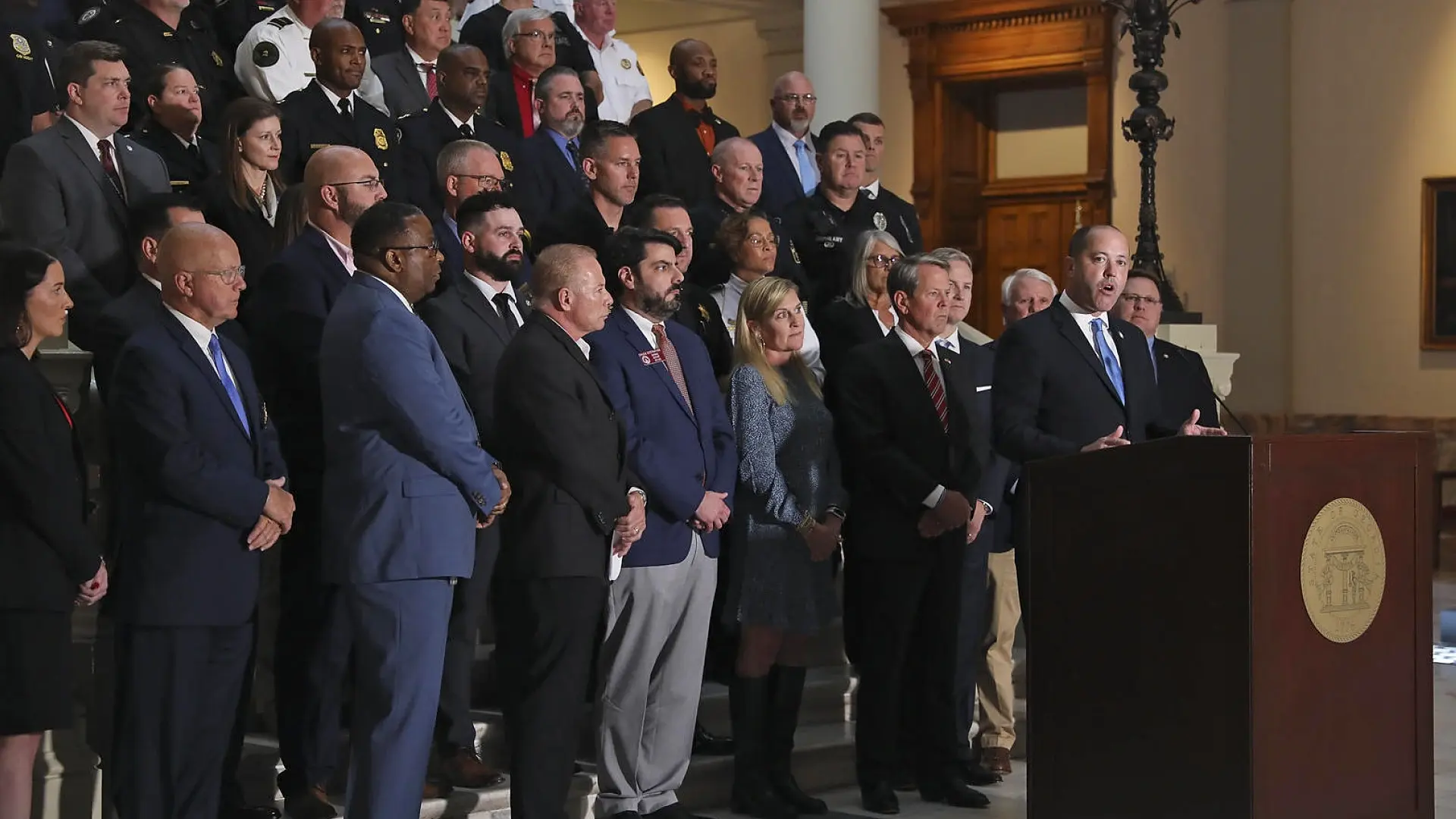 A press conference featuring officials and law enforcement personnel gathered at the Georgia Attorney General's Office, with a speaker addressing the audience from a podium.