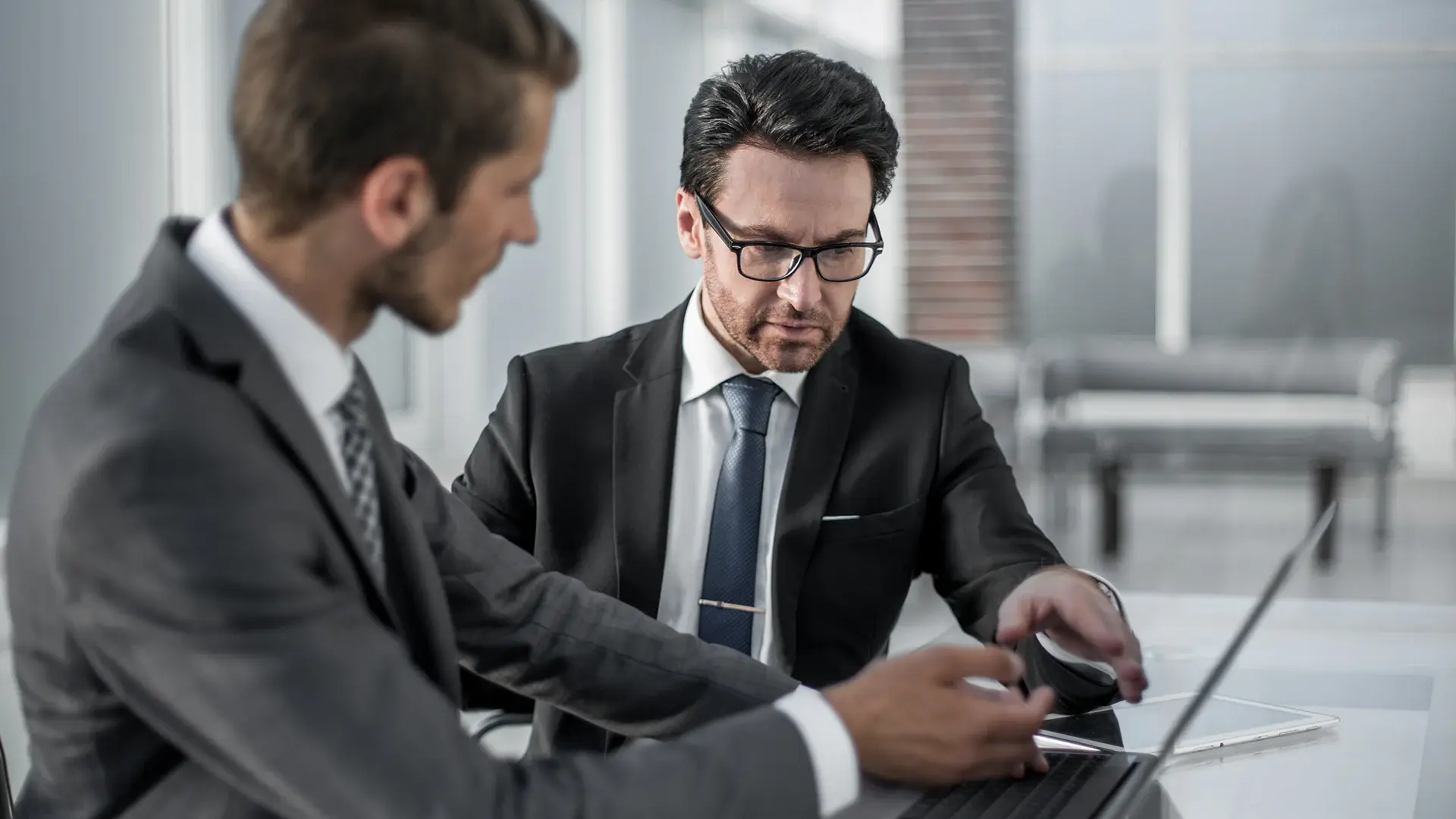 Two business professionals in suits reviewing information on a laptop during a meeting.